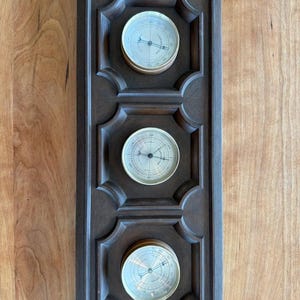 Puede incluir: Estación meteorológica de madera marrón oscuro para montaje en pared, con tres medidores circulares. Cada medidor tiene un bisel color latón y una esfera blanca con marcas negras y una aguja. El marco de madera tiene un diseño decorativo tallado.