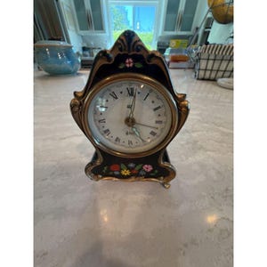 Puede incluir: Un reloj de sobremesa negro con bordes dorados y volutas ornamentadas. La esfera presenta números romanos, un emblema de corona y detalles florales pintados a mano en rojo, amarillo, azul y verde en la parte superior e inferior del marco.