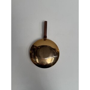 Puede incluir: Un objeto redondo de metal dorado con un asa de cuero marrón oscuro. El objeto tiene una superficie reflectante y parece ser una pieza decorativa o una bandeja pequeña. El asa está sujeta en la parte superior.