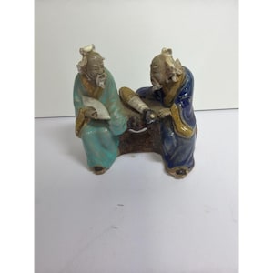 Op de afbeelding: Een keramieken beeldje met twee oudere figuren in traditionele gewaden. Eén figuur draagt een lichtblauwe jurk en houdt een waaier vast, terwijl de ander, in een donkerblauwe jurk, op een object lijkt te werken. De basis is bruin.