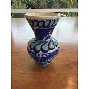 Puede incluir: Un jarrón de cerámica con base bulbosa y parte superior acampanada. El jarrón presenta un diseño floral azul y turquesa con detalles en blanco. El jarrón mide aproximadamente 15 cm de altura.