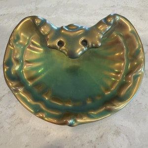 May include: A decorative, iridescent green and gold ceramic dish. The dish has a unique, scalloped edge and a raised, stylized design at the top with two small holes. The color shifts between green and gold, creating a shimmering effect.