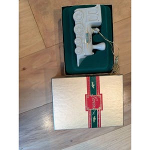 May include: A white ceramic train ornament with gold detailing, presented in a green velvet-lined box. The box is gold with red and green ribbon and the word "Lenox". A gold string is attached to the ornament.