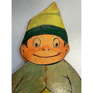 Puede incluir: Ilustración vintage de un personaje de dibujos animados sonriente con sombrero y chaqueta verdes. El personaje tiene piel naranja, ojos grandes y una nariz pequeña. El sombrero tiene una banda verde y la chaqueta tiene dos botones amarillos.