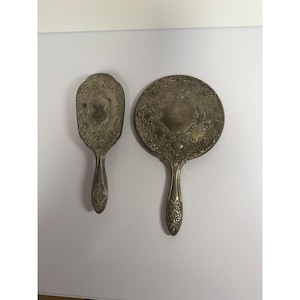 May include: A vintage silver-toned vanity set, featuring a hand mirror and a brush. Both items have ornate floral and scrollwork detailing. The mirror is round, and the brush is oval, each with a handle. The set is likely made of metal.