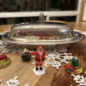 Puede incluir: Una mantequera plateada con tapa abovedada, rodeada de adornos navideños. Una pequeña figura de Papá Noel se encuentra sobre una base blanca. Otras decoraciones incluyen un pequeño cuenco verde con adornos y una vela.