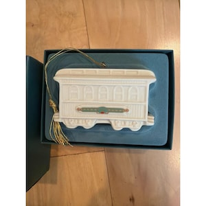May include: White ceramic train carriage ornament with gold hanging string. The ornament features detailed moulding and a decorative band with green and red accents. It is presented in a teal box with a teal interior.