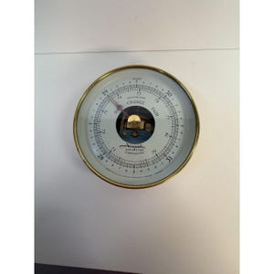 May include: A round Airguide barometer with a gold-toned metal frame and a white dial. The face displays dual scales in inches and centimeters, with the text "CHANGE" and "FAIR" printed in black. A red needle points to the current reading.