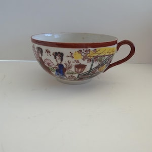 Peut inclure: Une tasse à thé en céramique blanche avec un bord et une anse marron. La tasse présente une illustration colorée de personnages asiatiques dans un cadre de jardin. L'œuvre comprend des personnages en vêtements bleus et marron, avec des accents jaunes et marron.