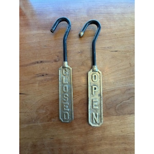 May include: Two vertical metal signs with black curved hooks at the top. One sign features the word CLOSED in raised gold-toned lettering on a rectangular plate, and the other features the word OPEN in identical gold-toned lettering and style.