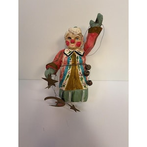 May include: A hand-painted wooden clown figurine with a red nose and cheeks, wearing a red and white patterned jacket with green accents. The figure has one arm raised and holds thin metal wires with dangling metal stars, a moon, and bells.