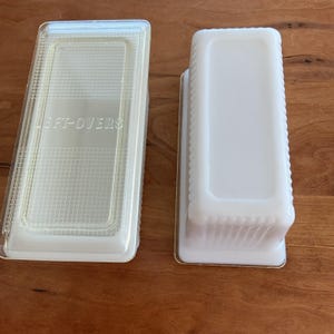 Op de afbeelding: Twee rechthoekige voedselcontainers in gebroken wit. Eén heeft een doorzichtig deksel met de tekst "LEFT-OVERS". De andere is open en effen gebroken wit. Beide staan op een houten oppervlak.