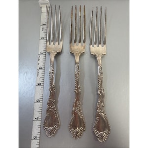 Op de afbeelding: Drie zilveren vorken met sierlijke, bloemmotief handvatten. De vorken zijn verticaal gerangschikt, met een meetlint ernaast. De vorken zijn ongeveer 20 cm lang.