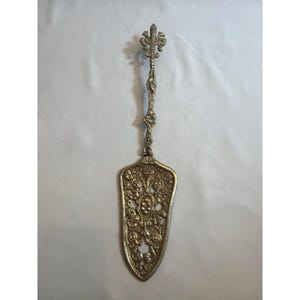 Puede incluir: Servidor de pasteles plateado con detalles florales y de volutas ornamentados. El mango presenta un diseño de flor de lis en la parte superior. La hoja del servidor tiene forma de escudo con patrones intrincados, ideal para servir pasteles.