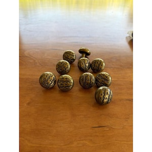 Peut inclure: Un ensemble de boutons de meubles de couleur bronze et un seul bouton en forme de champignon. Les boutons ont un motif texturé et gravé. La quincaillerie est disposée sur une surface en bois.