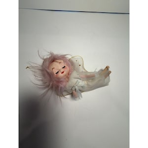 May include: A doll with light pink hair, closed eyes, and red lips, wearing a white dress with a small bow at the waist. The doll has sheer, gold-trimmed wings attached to the back and is positioned horizontally against a plain white background.