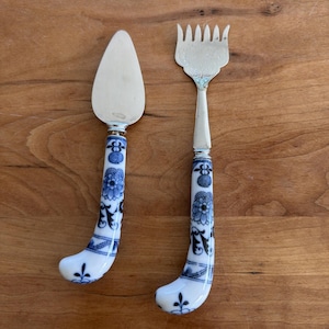 Puede incluir: Una espátula para pastel y un tenedor para servir plateados con mangos con estampado floral blanco y azul. Los mangos tienen un diseño curvo y son de porcelana. El tenedor para servir tiene múltiples púas.