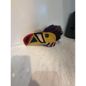 Può includere: Un oggetto decorativo a forma di uccello realizzato con fibre intrecciate in colori vivaci tra cui rosso, giallo, blu, bianco e nero. Il pezzo presenta un becco prominente, una testa strutturata con frange viola e marroni e una base.