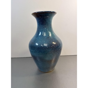 May include: A blue ceramic vase with a speckled finish. The vase has a flared rim and a rounded body. The color transitions from a dark blue at the base to a lighter blue towards the top, with a brown rim.
