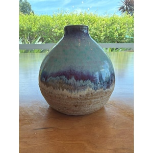 Könnte beinhalten: Eine handgefertigte Keramikvase mit rundem Körper und schmalem Hals. Die Vase hat einen Farbverlauf von Türkis, Lila und Beige. Die glänzende Oberfläche verleiht diesem dekorativen Stück einen Hauch von Eleganz. Ein einzigartiger Einrichtungsgegenstand.
