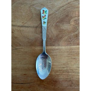 May include: A silver spoon with a detailed handle featuring the word "CANADA" and colorful maple leaves. The spoon's bowl has the word "ATHLETU" engraved. The spoon is set against a wooden surface.