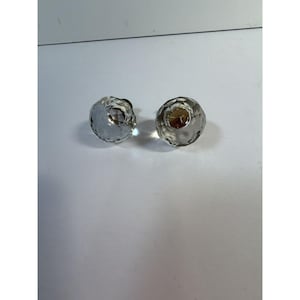 Pode incluir: Dois puxadores de vidro transparente facetado com formato redondo. Os puxadores têm uma superfície texturizada e uma base de metal prateado. Os puxadores são provavelmente para móveis ou uso decorativo.