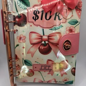 May include: A pink and clear savings challenge binder with a cherry and bow pattern. The binder has a pink clasp and the text "$10K". The words "savings challenge" are also visible.