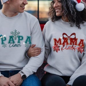 Puede incluir: Dos sudaderas grises claras con texto navideño. Una dice "PAPA Claus" en verde azulado con detalles de copo de nieve, y la otra dice "MAMA Claus" en rojo con detalles de lazo y flor de pascua. Llevadas por una pareja.