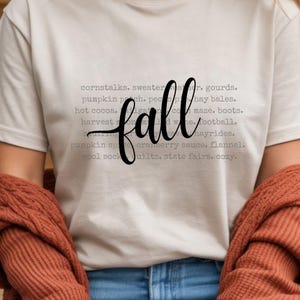 Puede incluir: Una camiseta color crema con la palabra "fall" en fuente negra. La camiseta presenta una lista de palabras con temática otoñal en fuente gris, que incluyen "tallos de maíz", "campo de calabazas" y "paseos en heno".