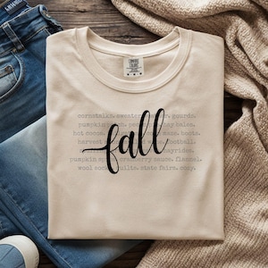 Op de afbeelding: Een crèmekleurig t-shirt met het woord "fall" in een zwart scriptlettertype. Het shirt heeft een lijst met herfstthemawoorden in een grijs lettertype, waaronder "maïsstengels", "pompoenveld" en "hooibalen".