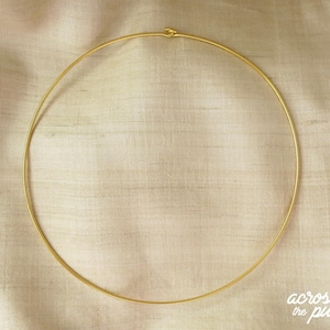 May include: A delicate, gold-colored choker necklace. The necklace is a simple, thin, circular design, with a small clasp at the top. The necklace is displayed on a light beige fabric.
