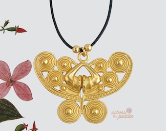 May include: A gold-toned pendant shaped like a stylized butterfly with intricate spiral and circular patterns, suspended from a thin black cord. The pendant features a symmetrical design with two larger bottom circles and four smaller top circles. The text "across the puddle" is visible in the lower right corner.