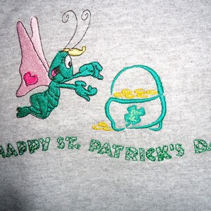 May include: A gray t-shirt with an embroidered design of a green bug with pink wings reaching for a pot of gold. The text "HAPPY ST. PATRICK'S DAY" is embroidered in green below the design.