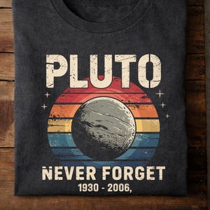 May include: Dark grey t-shirt featuring the word "PLUTO" in white block letters above a graphic of Pluto and the text "NEVER FORGET 1930-2006." The graphic has a retro sunset design with red, orange, yellow, and blue stripes.