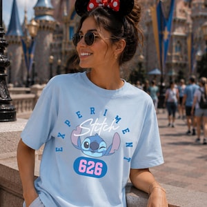 May include: Light blue t-shirt featuring a graphic of Stitch from Lilo & Stitch, with the text "Experiment" and "626". The shirt has a crew neck and short sleeves. The design is in shades of blue, pink, and white.