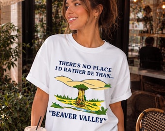 May include: White t-shirt with rolled sleeves featuring the text "THERE'S NO PLACE I'D RATHER BE THAN..." above a graphic of a tree and hills, with "BEAVER VALLEY" below. The shirt is worn by a person holding a beverage.