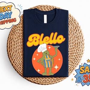 May include: A dark navy blue t-shirt featuring a graphic of a green character wearing a white chef hat and a multi-colored striped apron. The character holds a grey plate. Above the character, the word Blello is printed in orange and yellow.