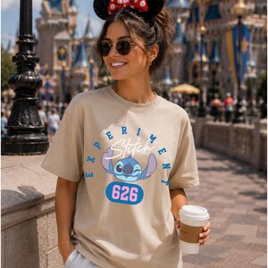 May include: A beige t-shirt featuring a graphic of the character Stitch from the movie Lilo & Stitch. The design includes the word "Experiment" and the number "626". The shirt is short-sleeved and has a crew neck.