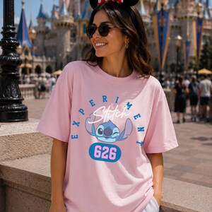 May include: A light pink t-shirt featuring a blue cartoon character with the text "EXPERIMENT Stitch" and the number "626". The shirt has short sleeves and a crew neck.