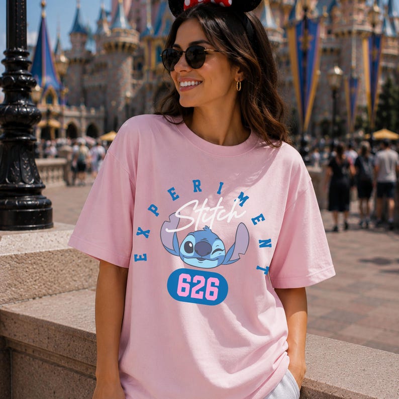 May include: A light pink t-shirt featuring a blue cartoon character with the text "EXPERIMENT Stitch" and the number "626". The shirt has short sleeves and a crew neck.