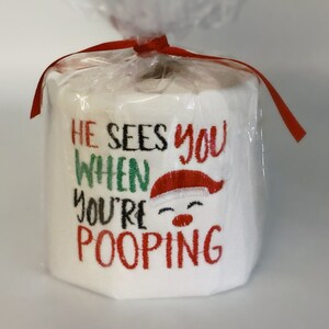 Puede incluir: Un rollo de papel higiénico blanco, envuelto en celofán transparente y atado con una cinta roja. El texto "HE SEES YOU WHEN YOU'RE POOPING" está bordado en rojo, verde y negro, con una cara de Papá Noel.