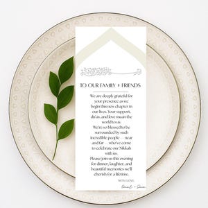 Puede incluir: Una mesa formal con dos platos blancos, una ramita de hojas verdes y una tarjeta blanca con texto. La tarjeta dice "A NUESTRA FAMILIA + AMIGOS" e incluye un mensaje sobre la celebración de un nuevo capítulo.