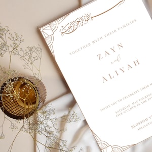 Puede incluir: Una invitación de boda blanca con caligrafía dorada y diseños geométricos. La invitación dice "Junto con sus familias, Zayn y Aliyah les invitan a celebrar su matrimonio el sábado 14 de mayo, a las cinco de la tarde. Blossom Venue, Arlington."