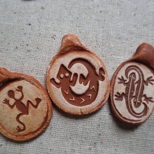May include: Three terracotta clay pendants with different lizard designs. Each pendant has a loop at the top for hanging.