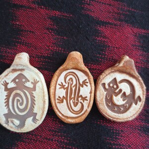 May include: Three ceramic pendants with brown lizard designs on a white background. The lizards are stylized and have different poses.