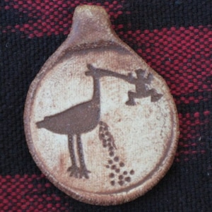 May include: A ceramic pendant with a brown and white design of a bird carrying a small figure in its beak. The bird is depicted with a long neck and legs, and the figure is holding a small object in its hand.