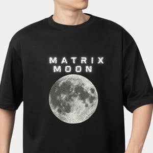 Może przedstawiać: Czarny t-shirt z białym napisem "MATRIX MOON" nad szczegółową białą ilustracją księżyca. Koszulka ma krótkie rękawy i okrągły dekolt.