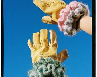 May include: Two yellow leather work gloves with different styles, one with a ruffled green knitted cuff and the other with a ruffled pink and gray knitted cuff. The gloves are held up against a bright blue sky.