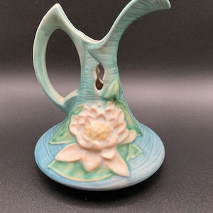 May include: A light blue ceramic pitcher with a handle and a spout. The pitcher features a raised design of a water lily with pink and green accents. The pitcher has a unique, open design near the handle.