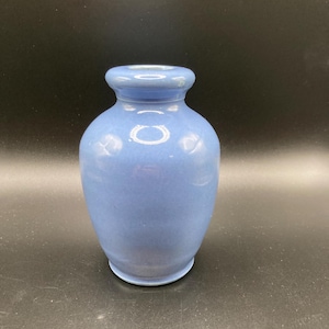 May include: A small, light blue ceramic vase with a rounded body and a narrow neck. The vase has a smooth, matte finish and a simple, classic design. The vase is approximately 6 inches tall.